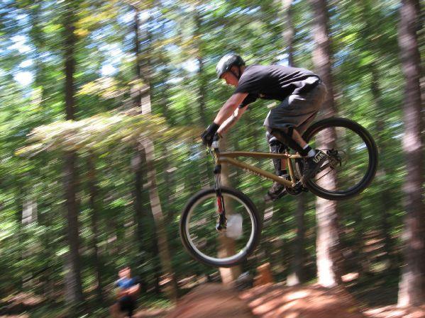 A mountain biker in a black shirt and helmet performs a jump over a dirt ramp in a wooded area, with trees and greenery in the background. The motion is captured in a dynamic way, emphasizing the thrill of the ride. Big Creek mountain bike trail.