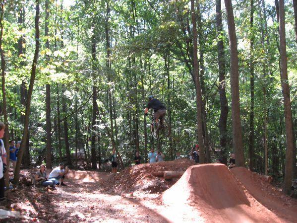A biker performing a jump over a dirt ramp in a wooded area, with spectators watching from the side. The scene captures the excitement of mountain biking in nature, surrounded by tall trees and a sunny atmosphere. Big Creek mountain bike trail.