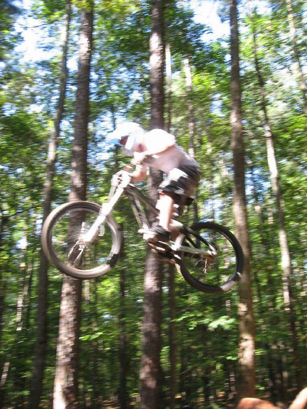 A mountain biker in mid-air, performing a jump on a wooded trail with tall trees in the background. The scene captures motion and excitement, showcasing the biker's skill and enthusiasm for the sport. Big Creek mountain bike trail.