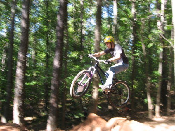 A person in a yellow helmet jumps off a dirt ramp while riding a mountain bike, surrounded by a dense forest with tall trees. The scene captures the thrill of biking in nature. Big Creek mountain bike trail.