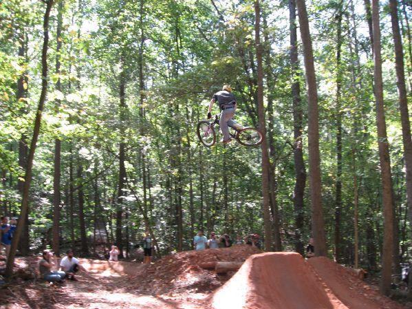 A biker performing a jump over a dirt ramp in a wooded area, surrounded by spectators watching the stunt. Big Creek mountain bike trail.