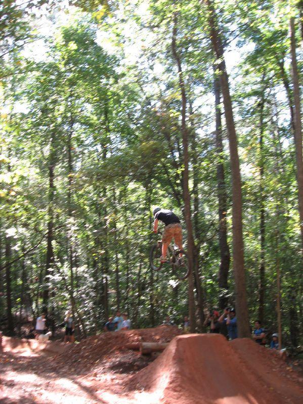 A cyclist performing a jump on a dirt bike in a forested area, with spectators watching in the background. The scene captures the excitement of outdoor biking and the natural surroundings. Big Creek mountain bike trail.