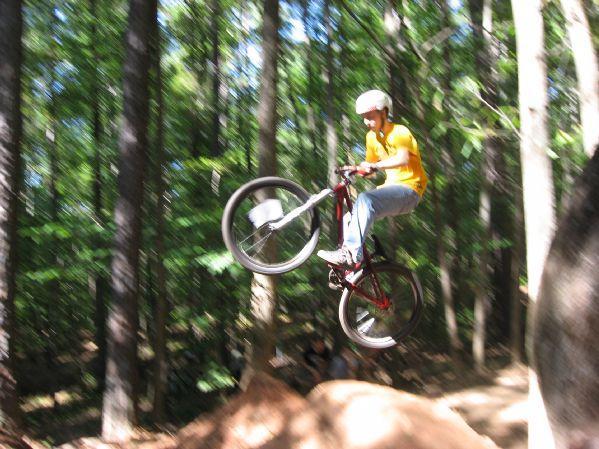 A person wearing a helmet and a yellow shirt performs a jump on a mountain bike over a dirt mound in a wooded area. The bike's front wheel is lifted off the ground as the rider is airborne, surrounded by trees and greenery. Big Creek mountain bike trail.