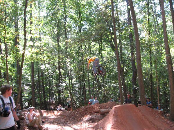 A BMX rider performing a jump over a dirt ramp in a wooded area, with spectators watching nearby. The scene is bright and sunny, showcasing the natural surroundings and the excitement of the event. Big Creek mountain bike trail.