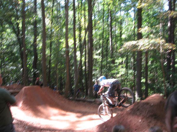 A mountain biker performing a jump over a dirt ramp in a wooded area, with spectators visible in the background. Tall trees and greenery surround the scene, creating a natural outdoor setting for biking. Big Creek mountain bike trail.