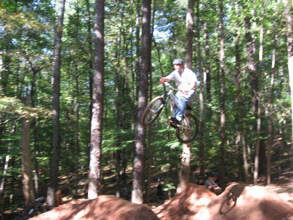 A person wearing a helmet and casual clothing is performing a jump on a mountain bike in a forested area. The background features tall trees and a dirt ramp, indicating a bike trail or park designed for cycling activities. Big Creek mountain bike trail.