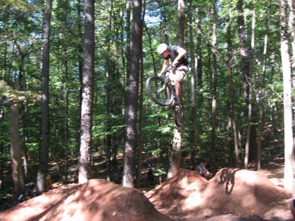 A mountain biker performing a jump over a dirt ramp in a wooded area, surrounded by tall trees and sunlight filtering through the leaves. Big Creek mountain bike trail.