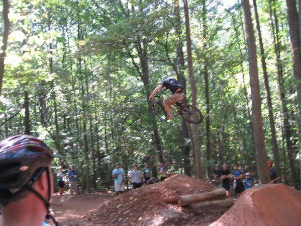 A person on a mountain bike performing a jump over a dirt ramp during a biking event, with a crowd of onlookers in the background. The setting is a wooded area with trees and sunlight filtering through the leaves. Big Creek mountain bike trail.