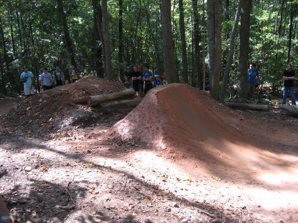 A dirt jump ramp in a forested area, surrounded by trees, with a group of people observing from the side. The sun filters through the leaves, casting dappled shadows on the ground and highlighting the smooth, packed dirt of the jump. Big Creek mountain bike trail.