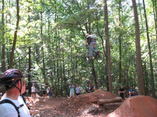 A cyclist performing a jump on a dirt ramp in a wooded area, surrounded by spectators watching the stunt. Sunlight filters through the trees, creating a vibrant outdoor setting. Big Creek mountain bike trail.