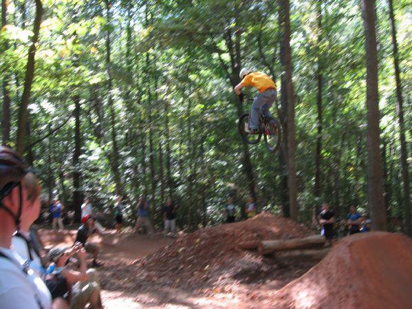 A cyclist performing a jump off a dirt ramp in a wooded area, surrounded by onlookers. The cyclist is wearing a yellow shirt and appears to be mid-air on a mountain bike, with trees and spectators visible in the background. Big Creek mountain bike trail.