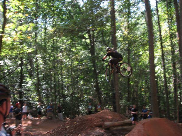 A mountain biker performing a jump in a forested area, with spectators watching from the side. The scene captures a moment of action with trees in the background and dirt ramps in the foreground. Big Creek mountain bike trail.