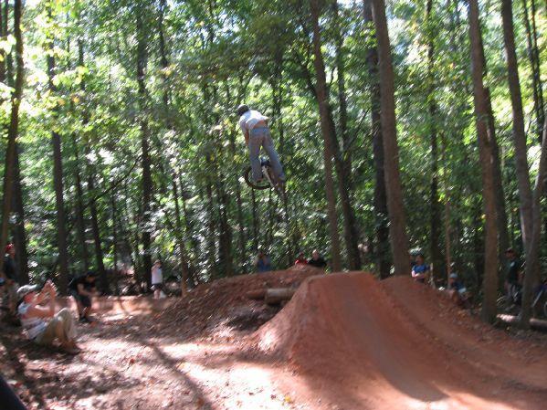 A cyclist performing a jump on a dirt bike ramp in a wooded area, surrounded by spectators seated and standing nearby, with sunlight filtering through the trees. Big Creek mountain bike trail.