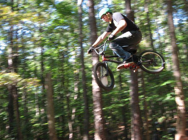 A cyclist performing a jump on a mountain bike in a forested area, surrounded by tall trees, with motion blur emphasizing the speed and height of the jump. The rider is wearing a helmet and protective gear. Big Creek mountain bike trail.