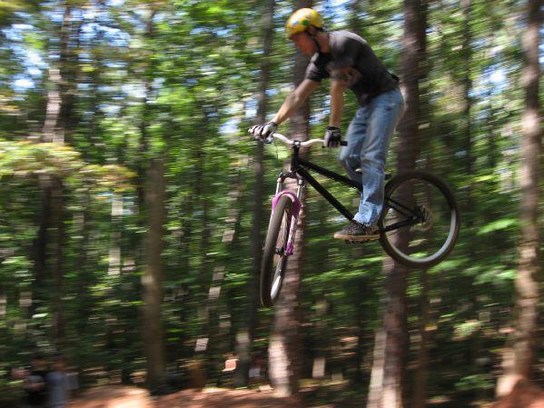 A person in a black shirt and blue jeans jumps a bicycle in mid-air, surrounded by tall trees in a forested area. The image captures a sense of motion, with a blurred background emphasizing the speed of the jump. The cyclist is wearing a yellow helmet and gloves, showcasing an adventurous outdoor activity. Big Creek mountain bike trail.