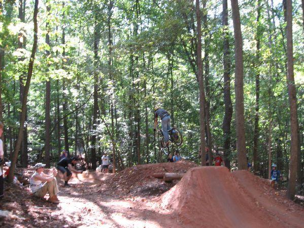 A BMX rider performing a jump over a dirt ramp in a wooded area, with spectators watching nearby. The scene captures the excitement of mountain biking and the beauty of nature. Big Creek mountain bike trail.