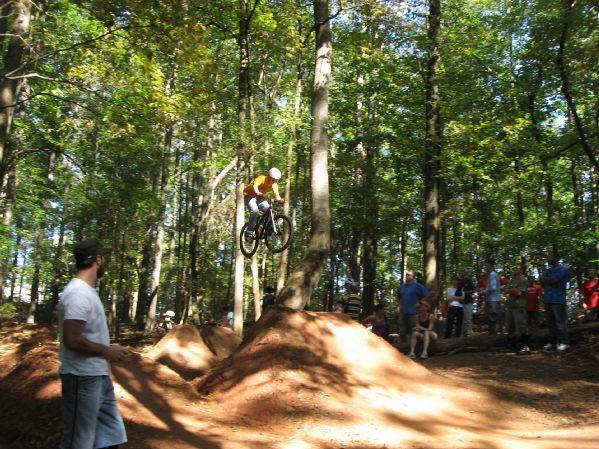 A biker in mid-air jumps over a dirt mound in a wooded area, while spectators watch from the sidelines. The sun filters through the trees, creating a vibrant outdoor scene. Big Creek mountain bike trail.