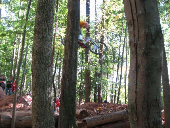A mountain biker performs a jump in a wooded area, surrounded by trees. Spectators watch from the side, with sunlight filtering through the leaves. The biker is mid-air, showcasing an action-packed moment against a natural backdrop. Big Creek mountain bike trail.