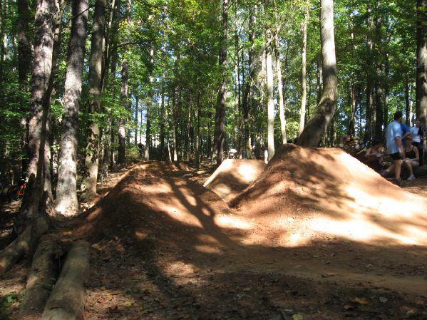 A dirt bike trail in a forested area featuring two mounds for jumps, with trees providing shade in the background. People can be seen observing the scene from the side. Big Creek mountain bike trail.