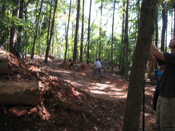 A mountain biker navigates a dirt trail through a wooded area, surrounded by tall trees and fallen logs. Spectators can be seen watching from the side, enjoying the outdoor activity in a sunlit forest. Big Creek mountain bike trail.