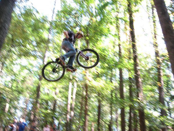 A mountain biker performing a jump on a trail surrounded by trees, with blurred motion indicating speed and excitement. Big Creek mountain bike trail.