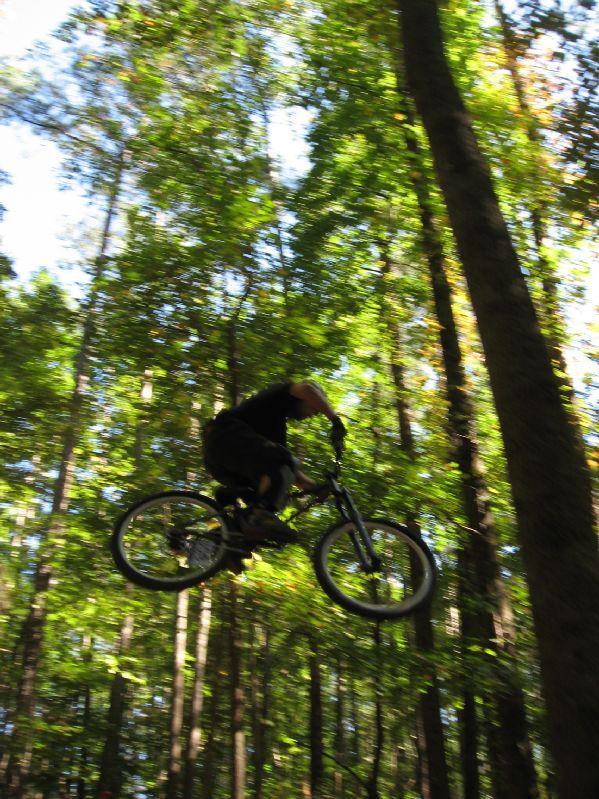 A mountain biker performs a跳跃 in a forested area, suspended in mid-air with trees and sunlight filtering through the leaves in the background. The image captures the excitement and motion of the sport. Big Creek mountain bike trail.
