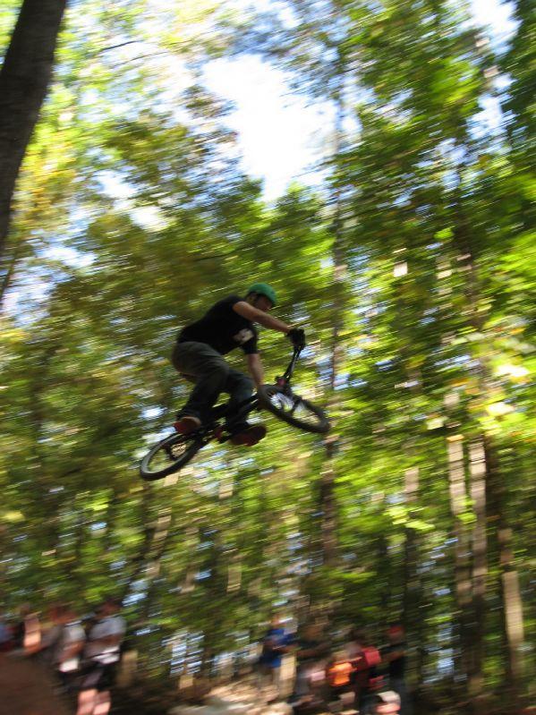 A BMX rider performs a jump in a forested area, surrounded by trees. The image captures the rider mid-air in a dynamic pose, creating a sense of motion. Spectators can be seen in the background, observing the performance. The scene is illuminated by natural light filtering through the leaves. Big Creek mountain bike trail.