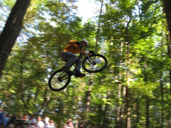 A cyclist wearing an orange shirt and helmet performs a jump on a mountain bike in a wooded area. The sun filters through the trees, creating a vibrant green backdrop, while spectators watch from the ground below. Big Creek mountain bike trail.