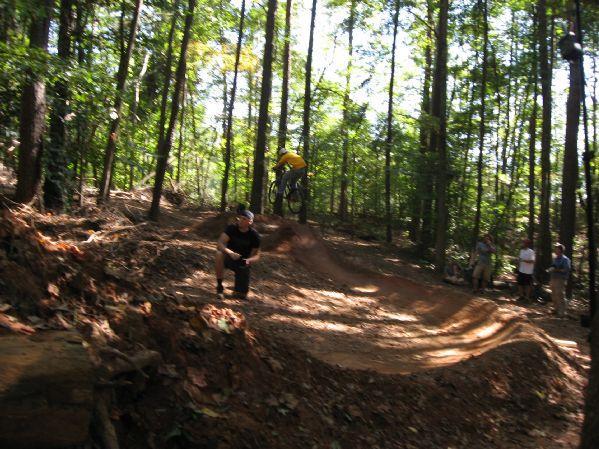 A mountain biker in a yellow shirt jumps off a dirt ramp in a wooded area, while a person in a black shirt crouches nearby. Several spectators watch from the side, surrounded by tall trees and sunlight filtering through the leaves. Big Creek mountain bike trail.