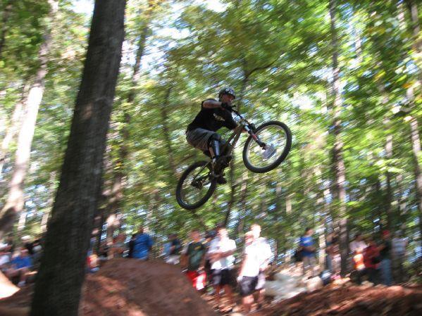 A mountain biker performing a jump in a wooded area, surrounded by spectators. The biker is airborne, showing dynamic motion, with a focus on the action of riding. Big Creek mountain bike trail.