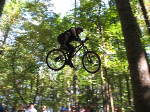 A mountain biker performing a jump over a dirt ramp in a wooded area, with spectators visible in the background watching the action. The cyclist is airborne, showcasing a dynamic pose amid tall trees and dappled sunlight filtering through the leaves. Big Creek mountain bike trail.