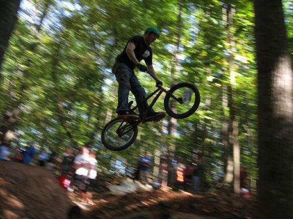 A BMX rider performs a jump over a dirt ramp in a wooded area, surrounded by spectators. The image captures the rider mid-air, with a blurred background emphasizing the motion and excitement of the stunt. The rider is wearing a helmet and casual clothing. Big Creek mountain bike trail.
