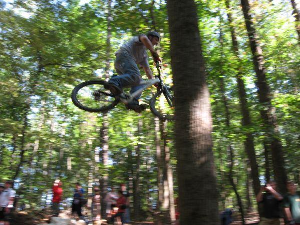 A mountain biker performs a jump over a tree in a forested area, surrounded by spectators watching the action. The scene captures a moment of excitement as the rider is airborne, showcasing the thrill of extreme sports. Big Creek mountain bike trail.