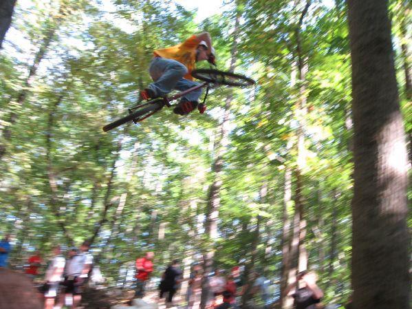 A mountain biker performing a jump over a rocky area in a wooded park, surrounded by spectators. The biker, wearing a yellow shirt and jeans, is mid-air, capturing the action and excitement of the moment. The background is filled with green trees and a blur of movement, suggesting speed. Big Creek mountain bike trail.