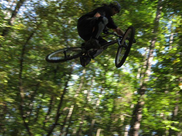 A cyclist performing a jump on a mountain bike, captured mid-air with a blurred background of tall trees and greenery. The rider is wearing a helmet and black clothing, showcasing a dynamic action pose against a natural outdoor setting. Big Creek mountain bike trail.