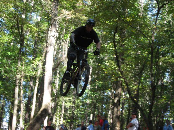 A mountain biker performing a jump over a wooded trail, surrounded by spectators watching the action. Sunlight filters through the trees, creating a vibrant and dynamic outdoor scene. Big Creek mountain bike trail.
