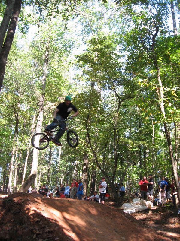 A BMX rider performing a jump in a wooded area, surrounded by spectators observing the action. The rider is airborne above a dirt ramp, showcasing a dynamic stunt amidst lush green trees and a crowd in the background. Big Creek mountain bike trail.