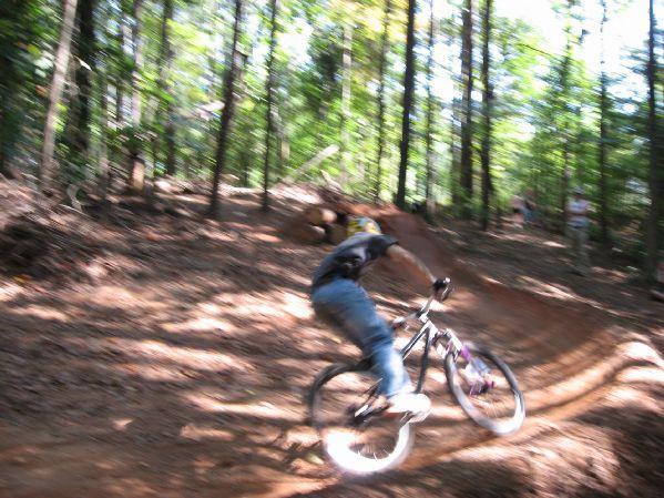 A mountain biker in motion navigating a dirt trail through a wooded area, surrounded by trees and dappled sunlight. The rider is leaning toward one side, showcasing the dynamic action of off-road biking. Big Creek mountain bike trail.