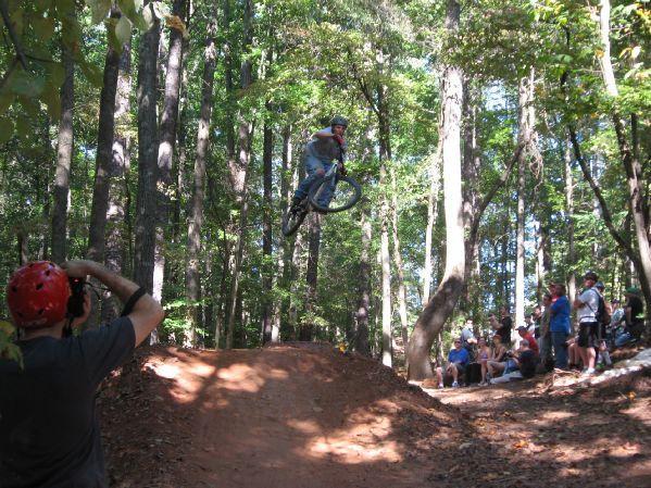 A BMX rider performs a jump over a dirt ramp in a wooded park, while a spectator with a camera captures the action. A small crowd watches from the side, surrounded by tall trees and dappled sunlight filtering through the leaves. Big Creek mountain bike trail.