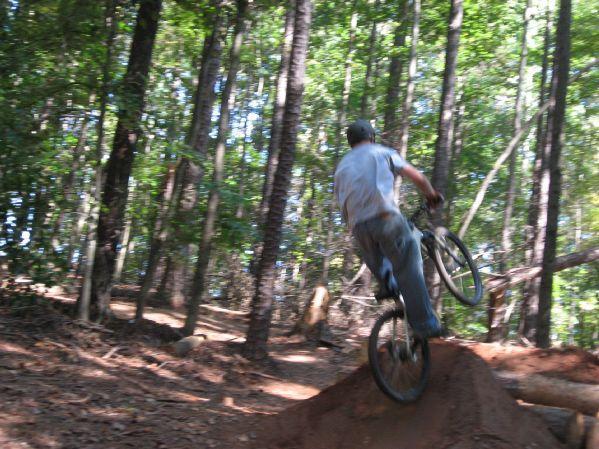 A person riding a mountain bike jumps off a dirt ramp in a wooded area, surrounded by tall trees and a natural trail. The rider is wearing a gray shirt and a cap, with the bike elevated off the ground as they perform the jump. Big Creek mountain bike trail.