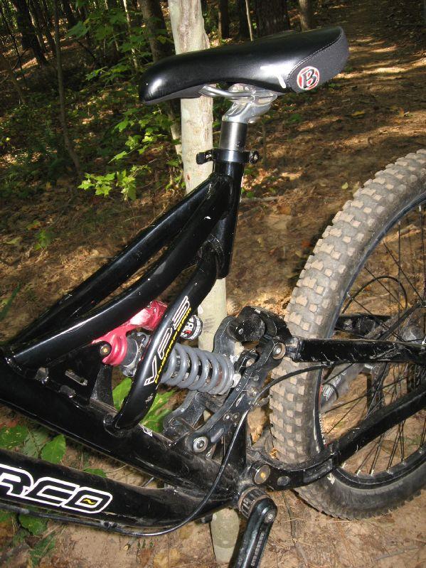 Close-up view of a mountain bike frame featuring a black and red suspension system, with a visible seat and tire. The bike is positioned next to a tree in a wooded area, showcasing the natural surroundings with leafy vegetation and dirt trail. Big Creek mountain bike trail.