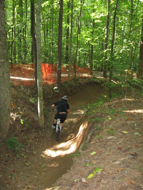 A person riding a mountain bike down a dirt trail in a wooded area, with green trees surrounding the path and orange safety fencing in the background. Big Creek mountain bike trail.