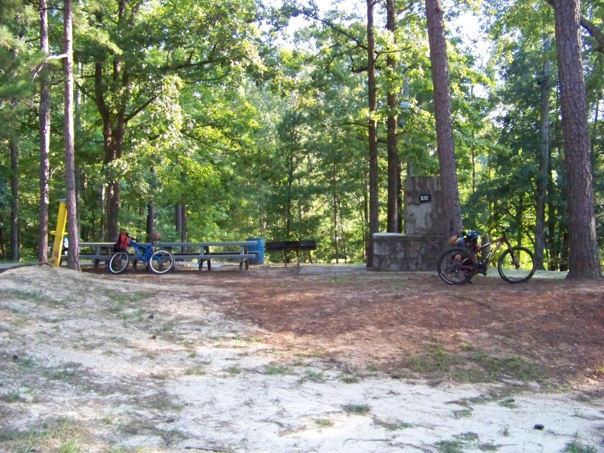A peaceful outdoor scene featuring two bicycles parked near a picnic area in a forest. Tall trees surround a gravel clearing with a picnic table and a stone marker labeled "612." The setting conveys a sense of tranquility and outdoor adventure. Flat Rock Park mountain bike trail.