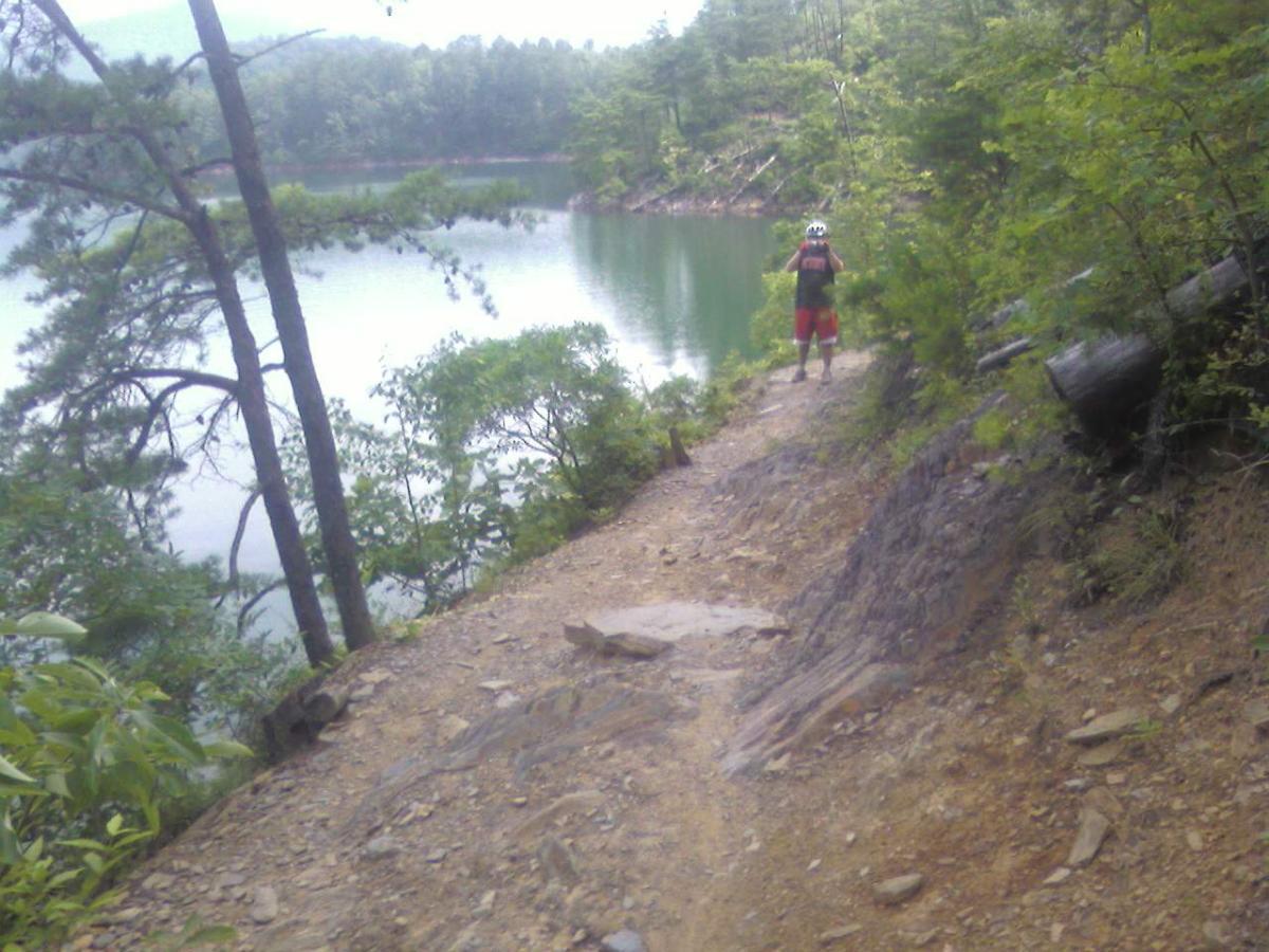 A person wearing a helmet and athletic attire stands on a rocky trail beside a calm lake, surrounded by lush green trees and foliage. The scenery features a peaceful natural landscape with water reflecting the surrounding trees. Tsali Right Loop mountain bike trail.