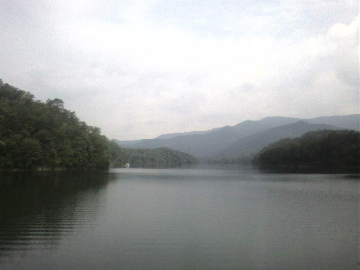 A serene view of a calm lake surrounded by lush greenery and distant mountains, under a cloudy sky. The water reflects the landscape, creating a peaceful and tranquil atmosphere. Tsali Right Loop mountain bike trail.