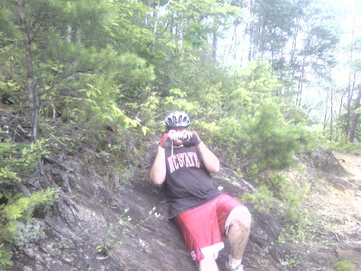 A person wearing a bicycle helmet and a black shirt is sitting on a rocky surface in a wooded area, looking at the camera while holding a camera. Their shorts are muddy, suggesting recent activity, and they are surrounded by greenery. Tsali Right Loop mountain bike trail.