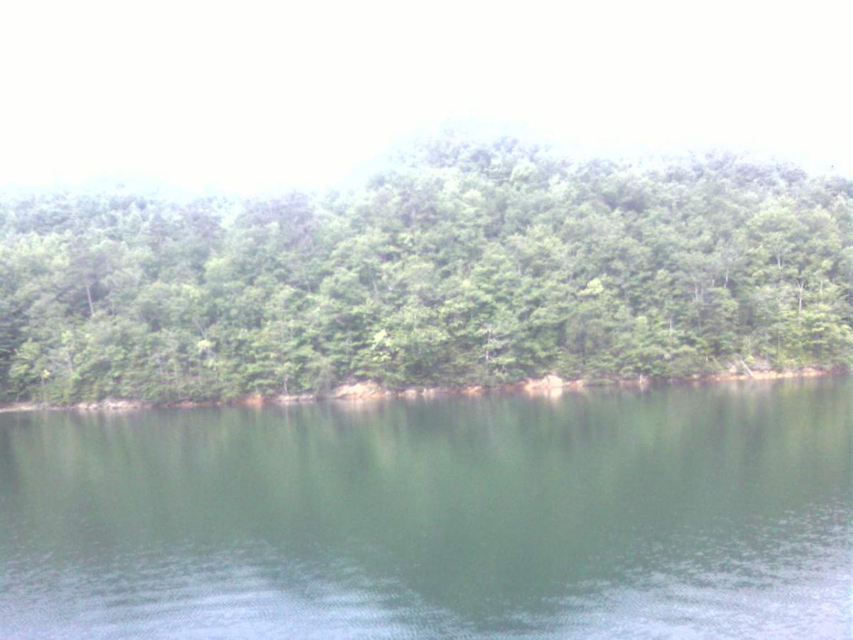 A serene landscape featuring a calm lake reflecting surrounding greenery, with a dense tree line along the shore and mist obscuring the distant hills. The water appears still, creating a peaceful atmosphere. Tsali Right Loop mountain bike trail.
