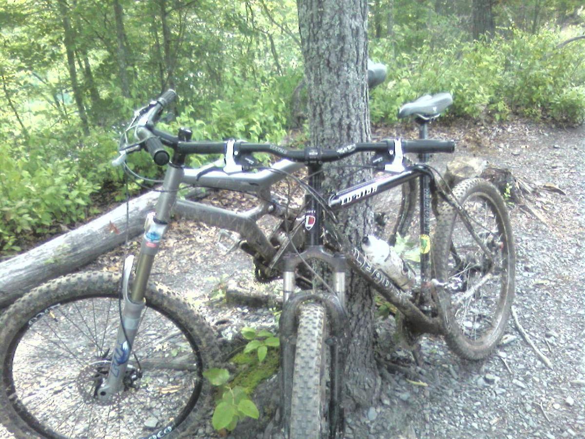 A close-up view of a mountain bike leaning against a tree in a wooded area. The bike is partially covered in dirt, indicating recent use on a trail, with a water bottle attached to the frame and greenery surrounding the scene. Tsali Right Loop mountain bike trail.