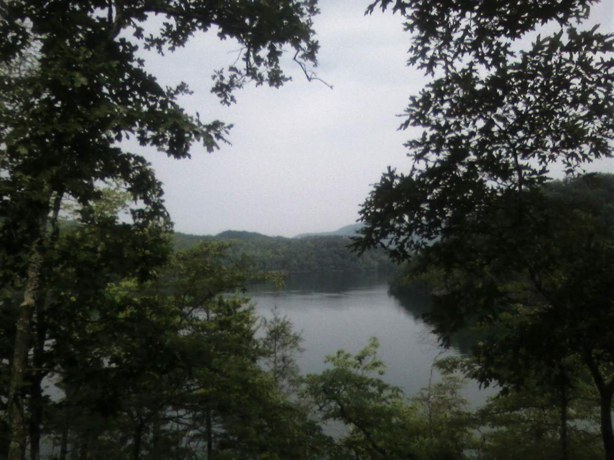 Serene view of a calm lake framed by lush green trees, with misty mountains in the background under a cloudy sky. Tsali Right Loop mountain bike trail.