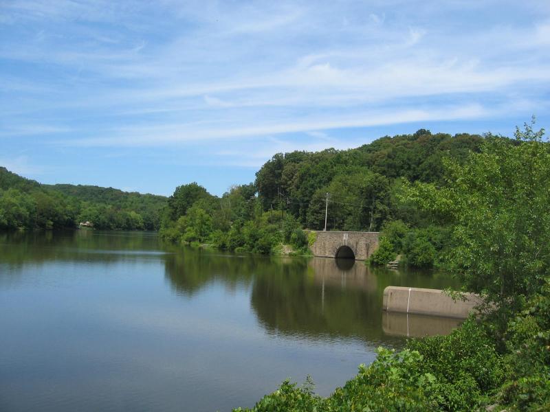 Scenic view of a calm river surrounded by lush green trees under a clear blue sky, featuring a stone bridge with an archway reflected in the water. North Park mountain bike trail.